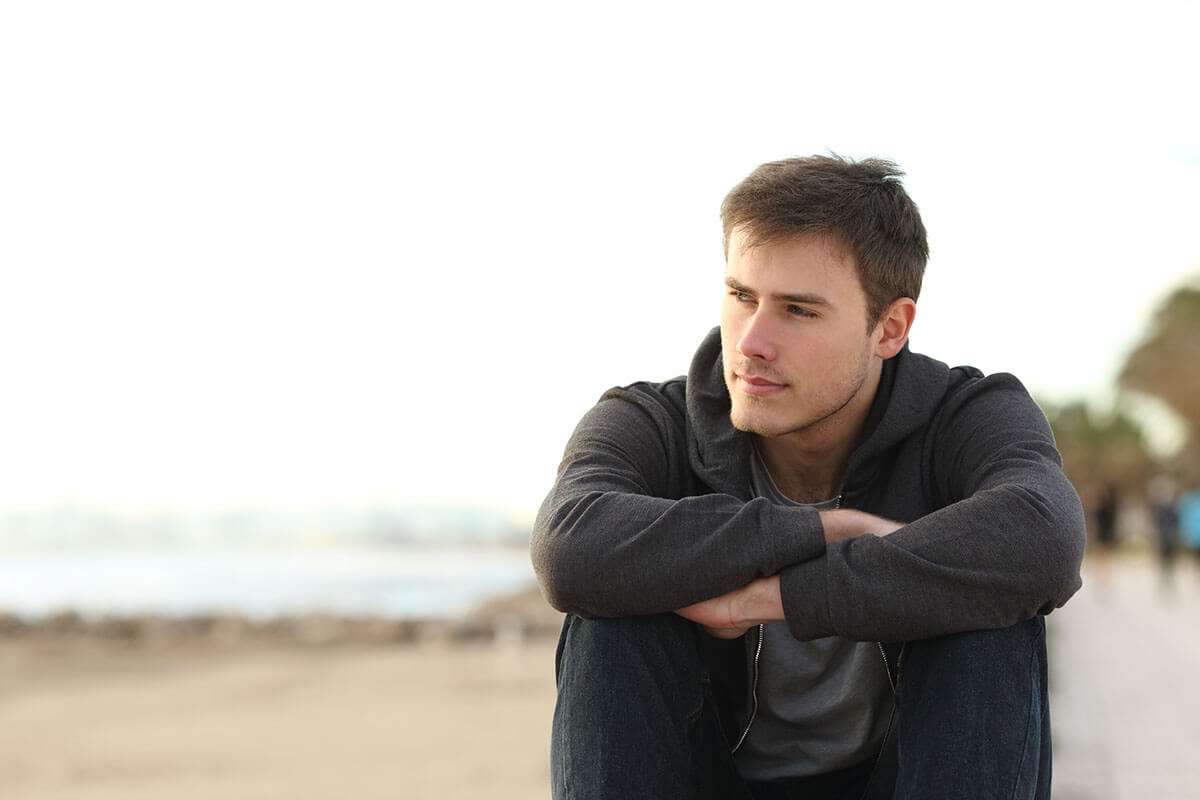 man sits on beach in a hoodie as he realizes the signs of depression in men